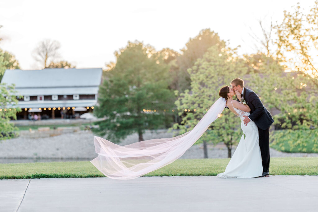 Bride and Groom dip kissing with veil flying at sunset with water behind them